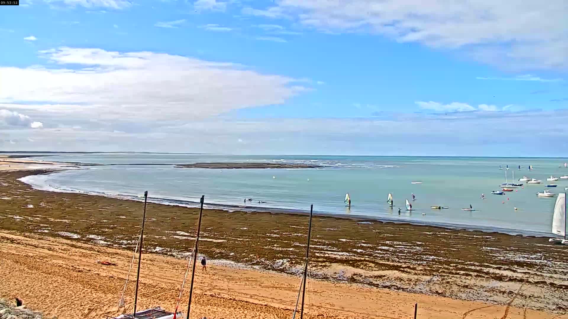 A sandy beach with seaweed exposed at low tide stretches toward a calm, light-green ocean where several sailboats and windsurfers are visible under a partly cloudy sky.