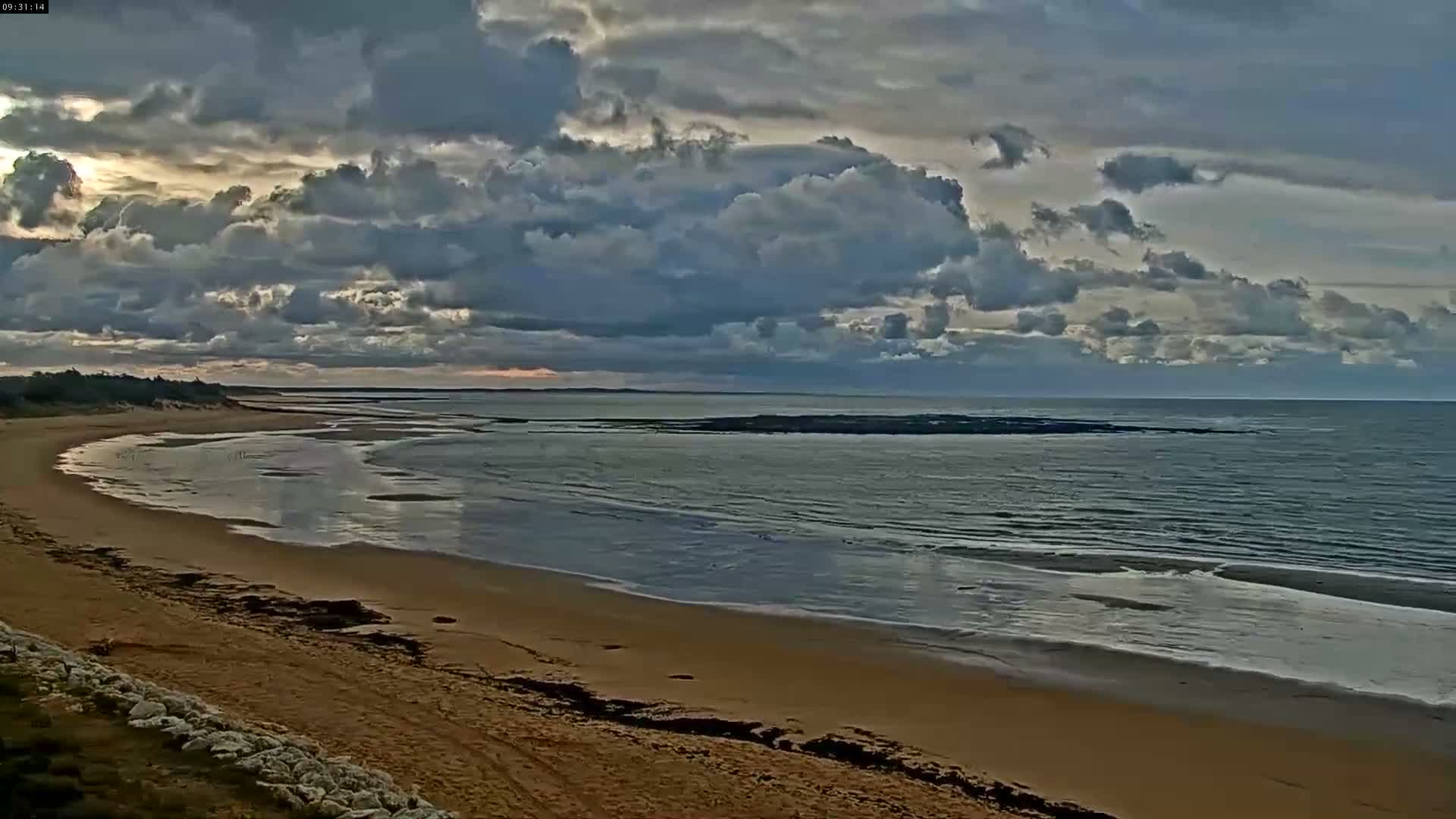 A vast sandy beach curves along a calm ocean under a heavily clouded sky with hints of light breaking through.