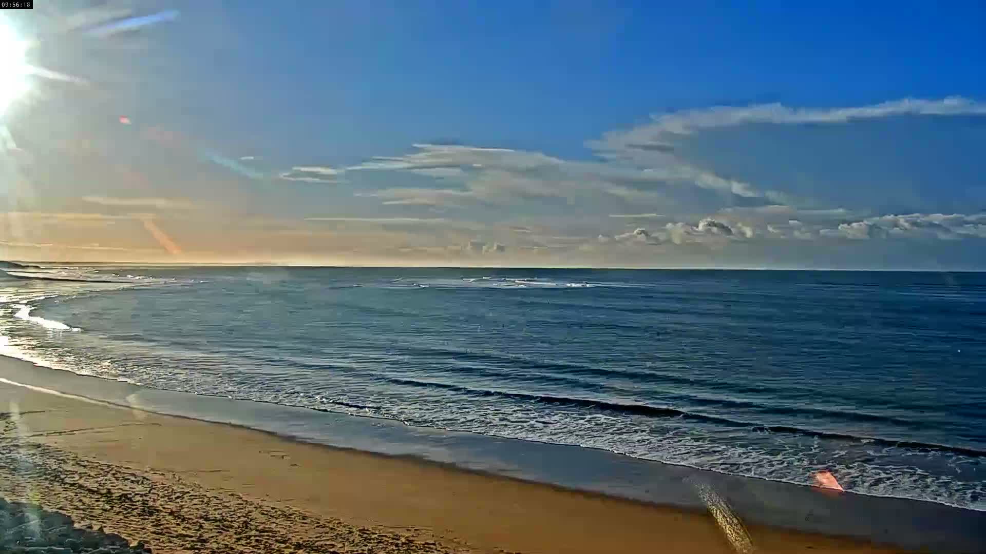 A bright, sunny beach scene features a wide expanse of golden sand meeting gentle ocean waves under a partly cloudy blue sky, with the sun low on the horizon.