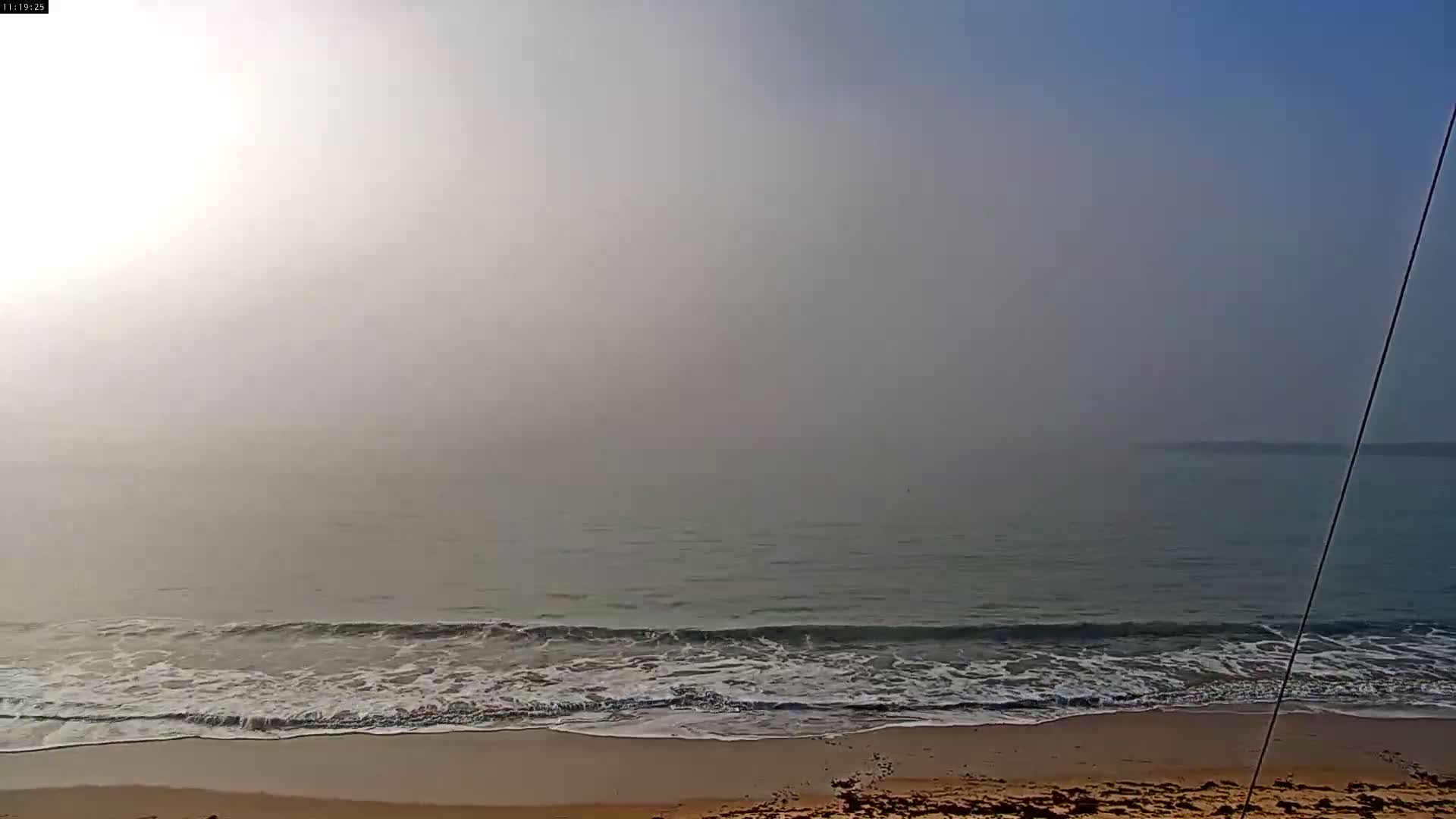 A bright, sunny beach scene features a wide expanse of golden sand meeting gentle ocean waves under a partly cloudy blue sky, with the sun low on the horizon.