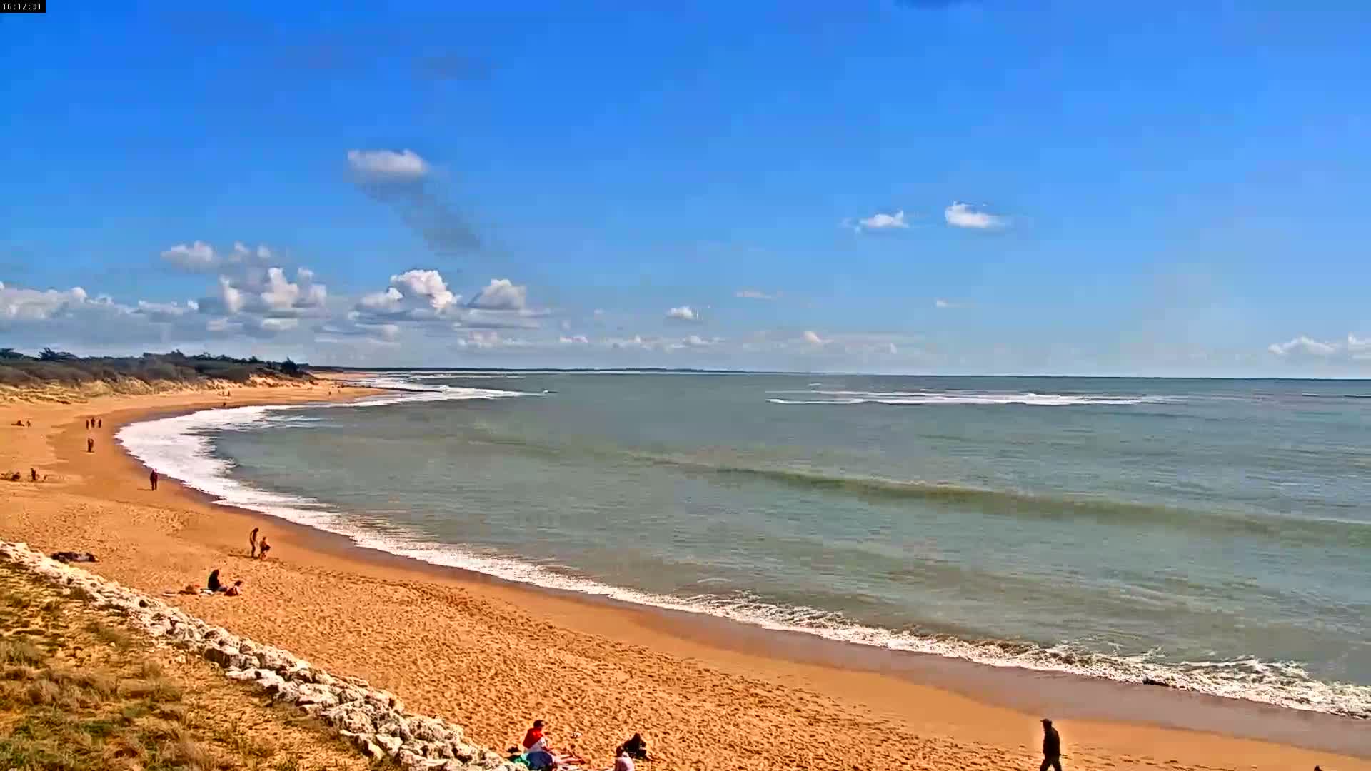 A bright, sunny beach scene features a wide expanse of golden sand meeting gentle ocean waves under a partly cloudy blue sky, with the sun low on the horizon.
