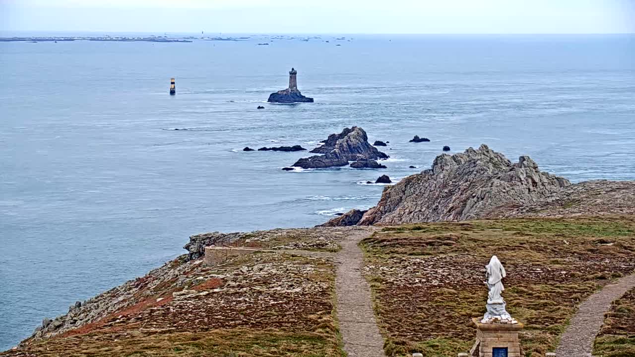 A rocky coastal scene with a distant lighthouse and a statue overlooking a path on a cloudy day.
