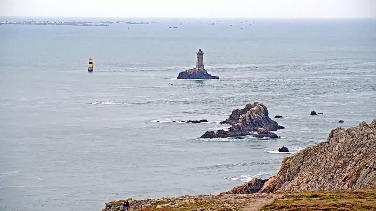 A coastal scene on a cloudy day shows a lighthouse on a small island, dark rocky outcrops, a distant landmass, and two people standing on a cliff.