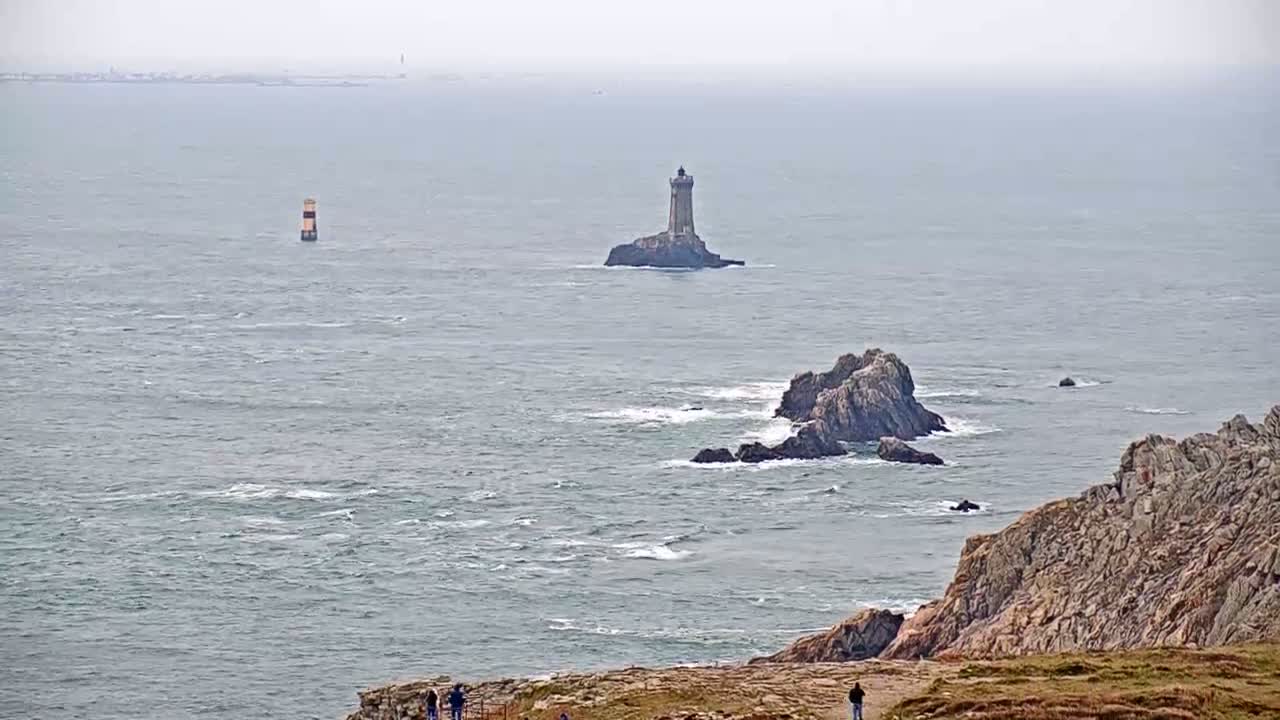 A rocky coastline with a lighthouse on a small island in the distance, under an overcast sky with a slightly choppy sea.