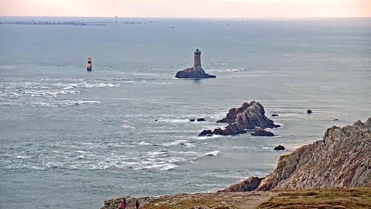 A rocky coastline with a lighthouse on a small island in a choppy sea under a mostly cloudy sky.