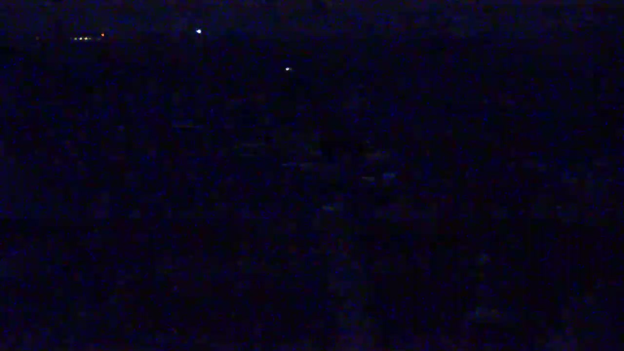 A dark, nighttime scene shows a large, indistinct mass possibly representing a crowd or field, with a few faint lights visible in the distance.