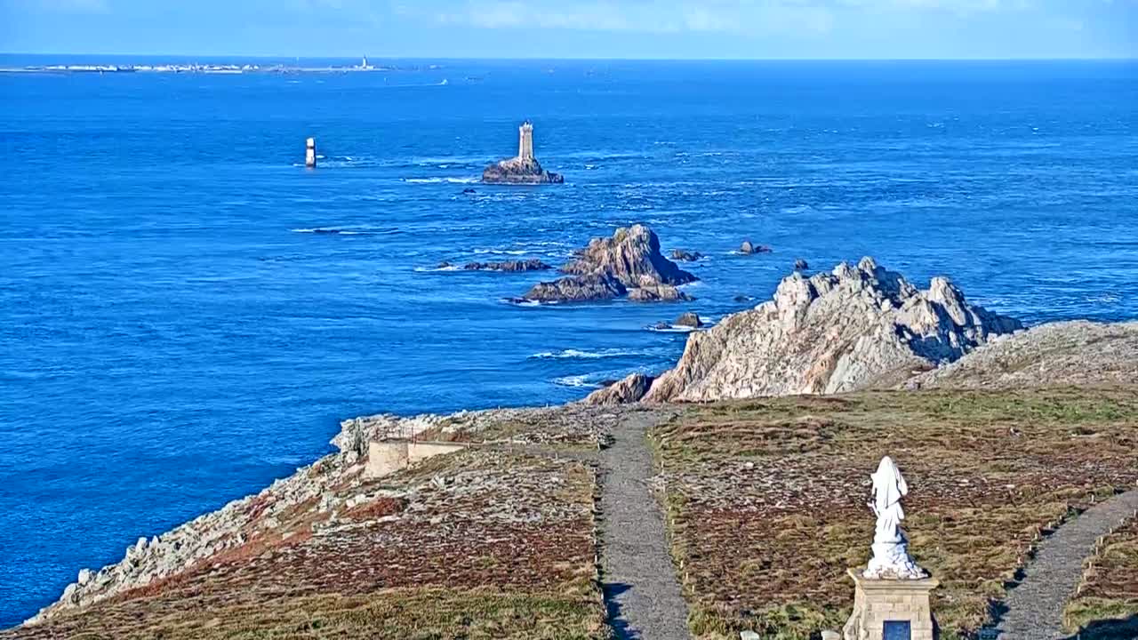 A coastal scene on a sunny day shows a rocky shoreline with a statue, a path, and distant islands and lighthouses in a deep blue ocean.