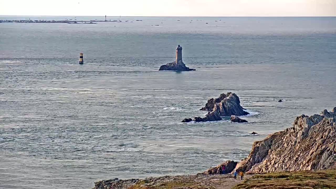 A rocky coastline overlooks a choppy sea with a distant lighthouse and a smaller marker buoy visible, under a mostly clear sky.