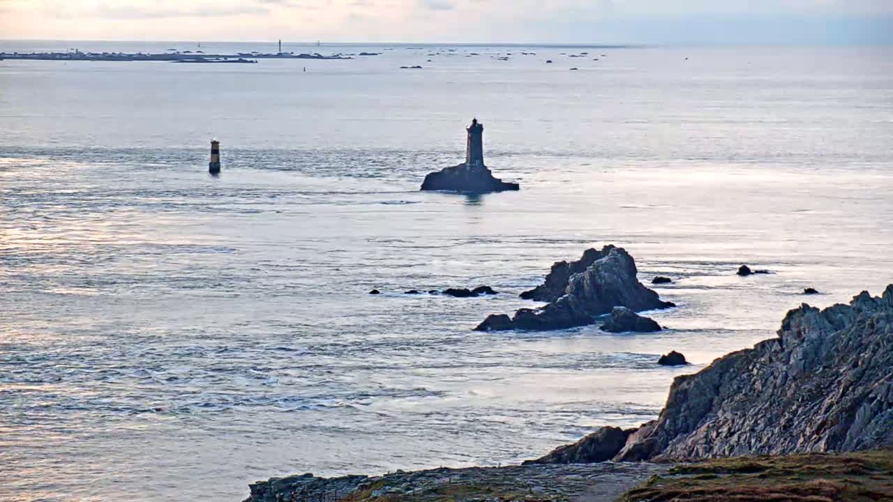 A rocky coastline with a lighthouse on a small island in the distance, under an overcast sky, with choppy water.