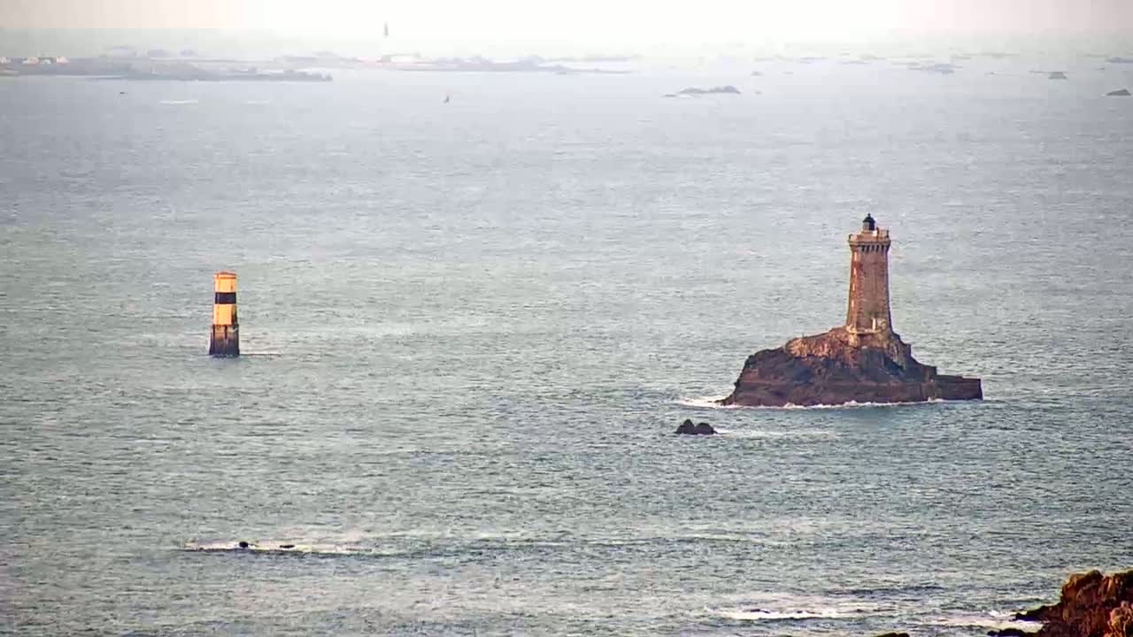 A tall lighthouse sits on a small, rocky island in a calm, gray sea, with a smaller, striped lighthouse visible in the distance.