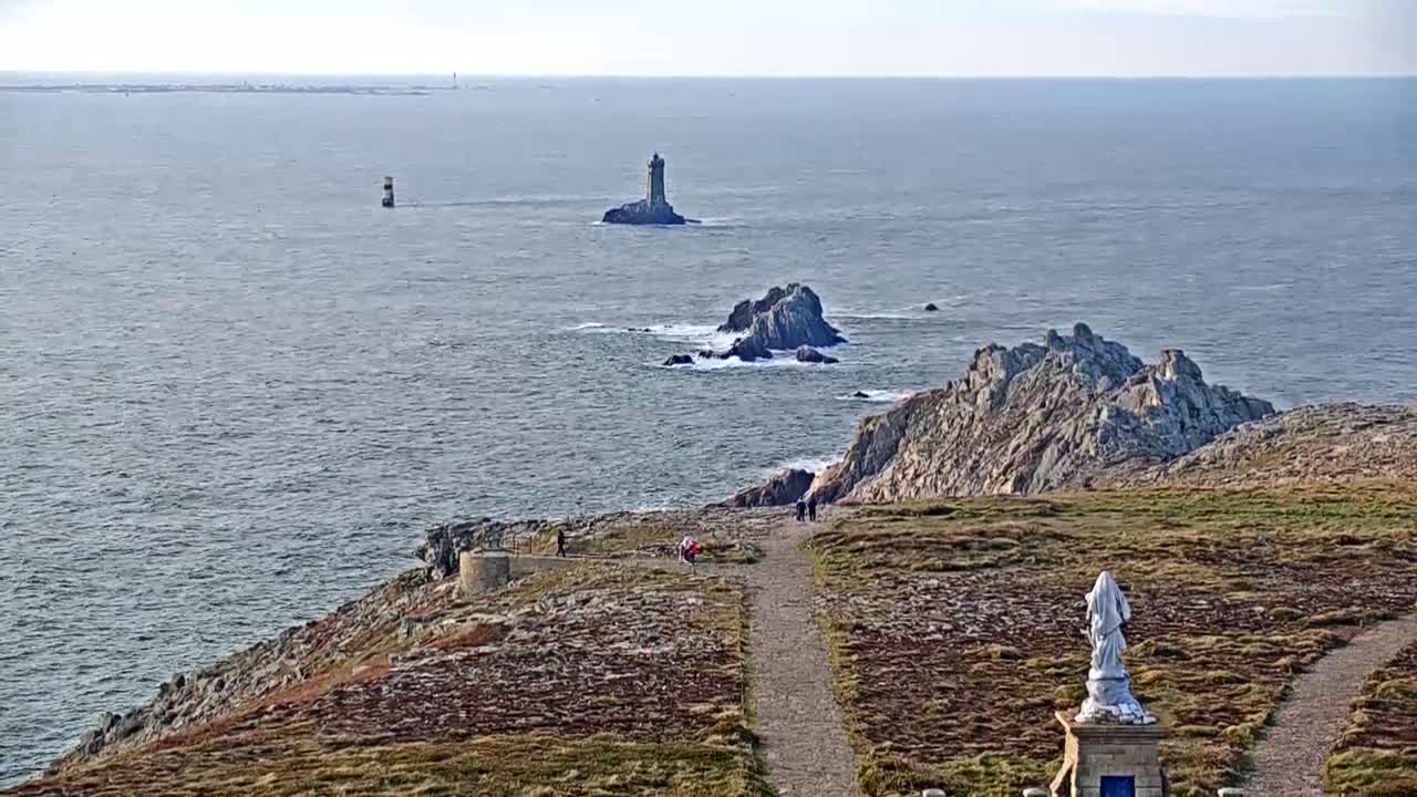 A coastal scene on a partly sunny day shows a rocky outcrop with a statue and walking path overlooking the ocean where two lighthouses and other rocky islands are visible in the distance.