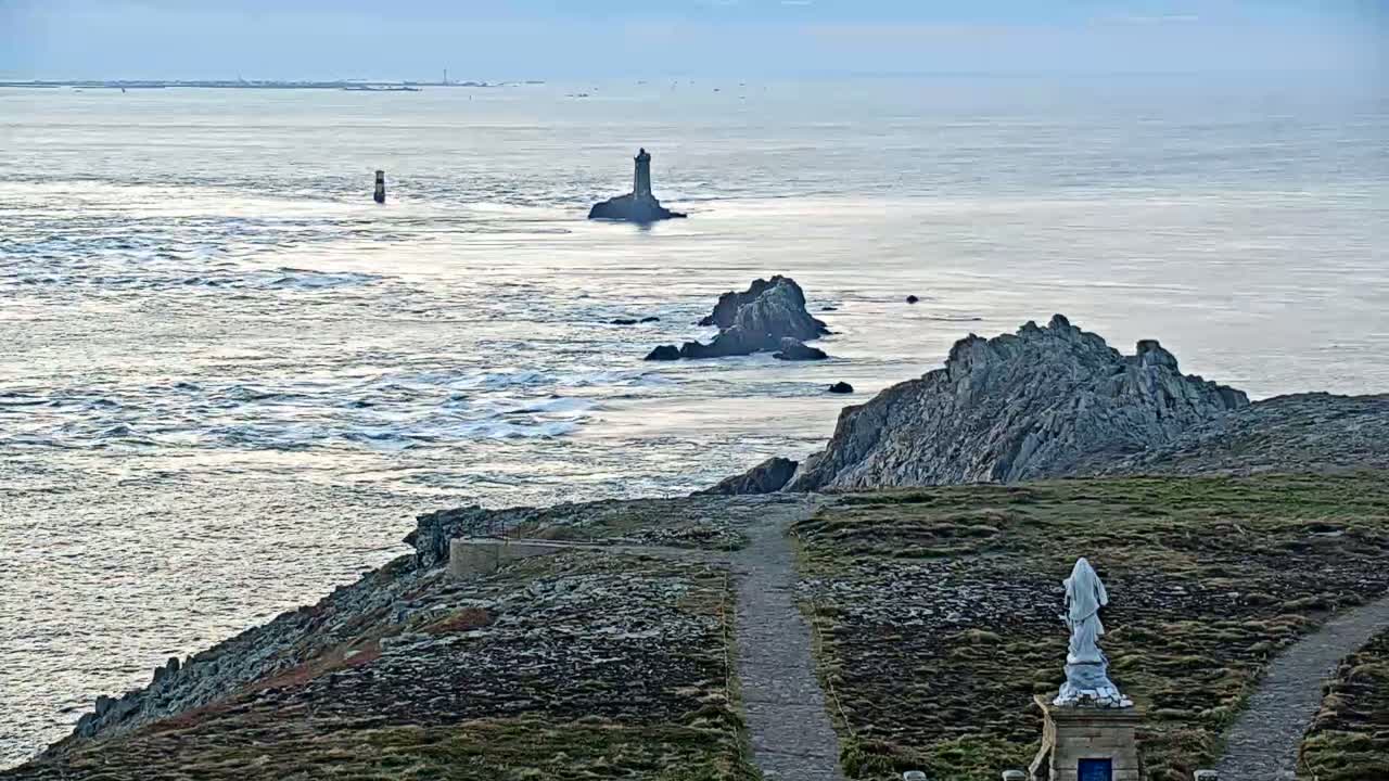 A statue stands on a rocky cliff overlooking a calm sea with two lighthouses and several rocky outcrops visible in the distance under a pale sky.