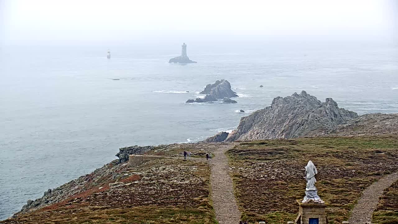 A statue stands on a rocky, grassy coastal path overlooking a foggy ocean with several islands and a distant lighthouse.