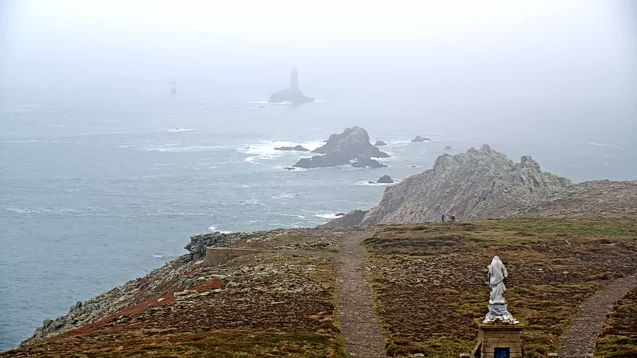A statue stands on a grassy coastal path overlooking a foggy ocean with rocky outcrops and distant lighthouses.