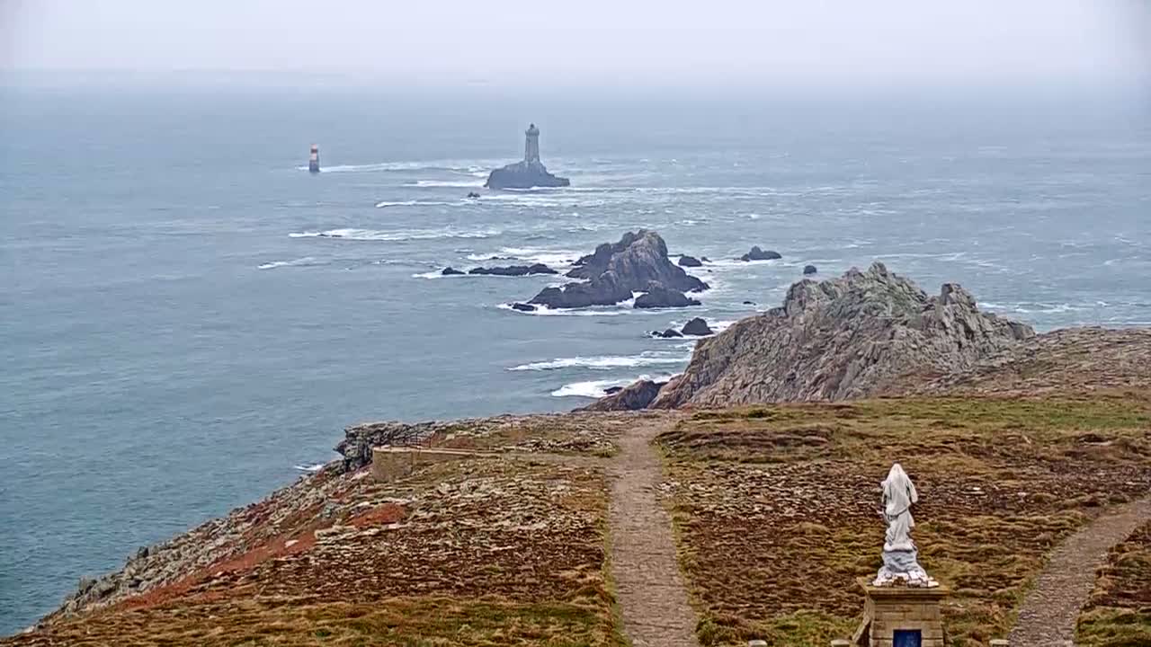 A statue stands on a grassy cliff overlooking a wavy ocean with two distant lighthouses and rocky outcrops under an overcast sky.