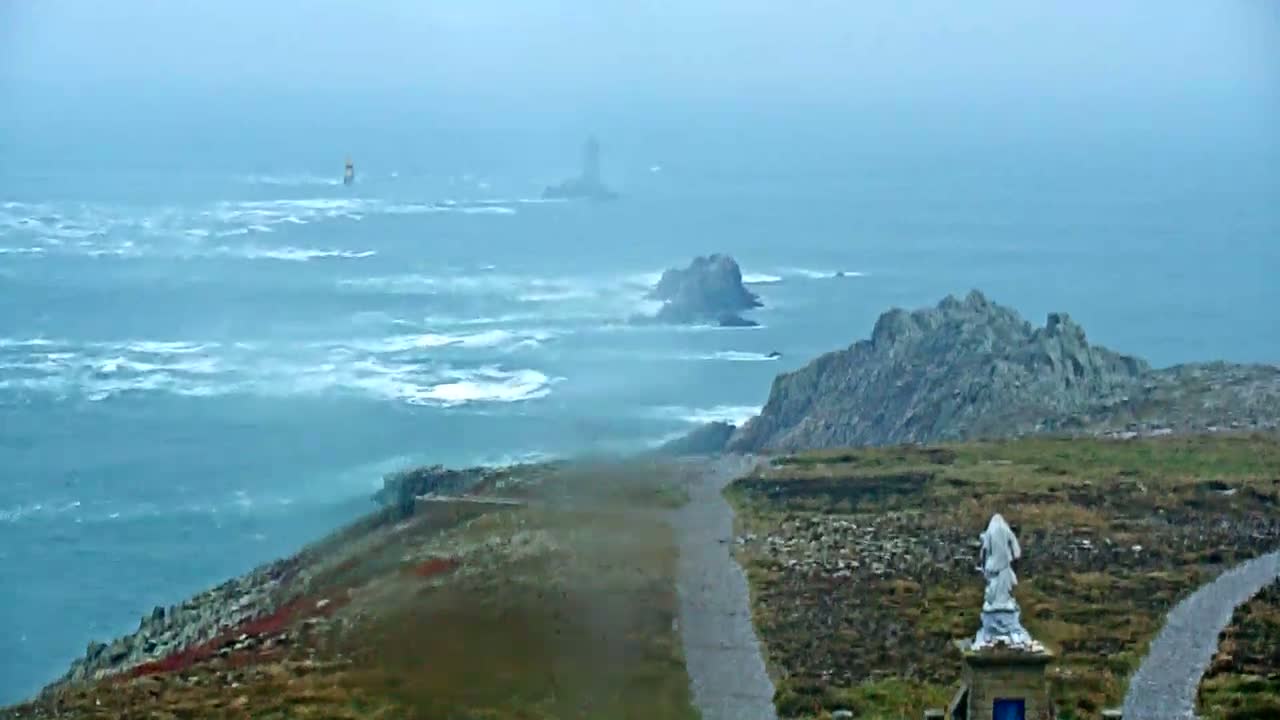 A statue stands on a coastal path overlooking a stormy sea with distant rocky outcrops and a lighthouse.
