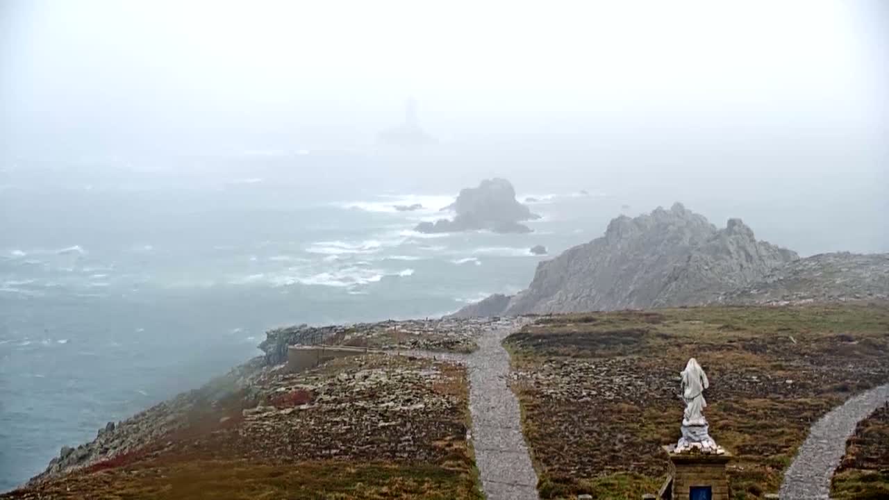 A statue stands on a rocky path overlooking a foggy, turbulent ocean with distant, shrouded cliffs.