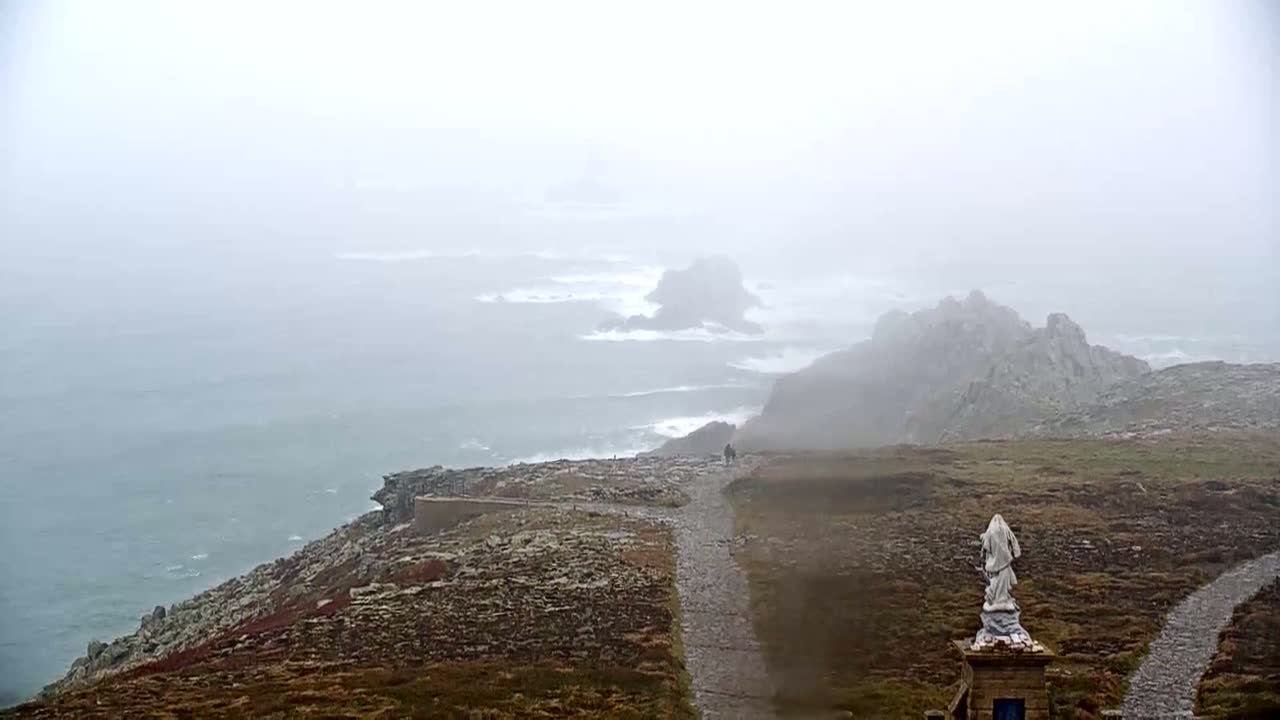 A foggy coastal scene shows a statue on a path overlooking a rocky, ocean shoreline with waves crashing against the rocks.