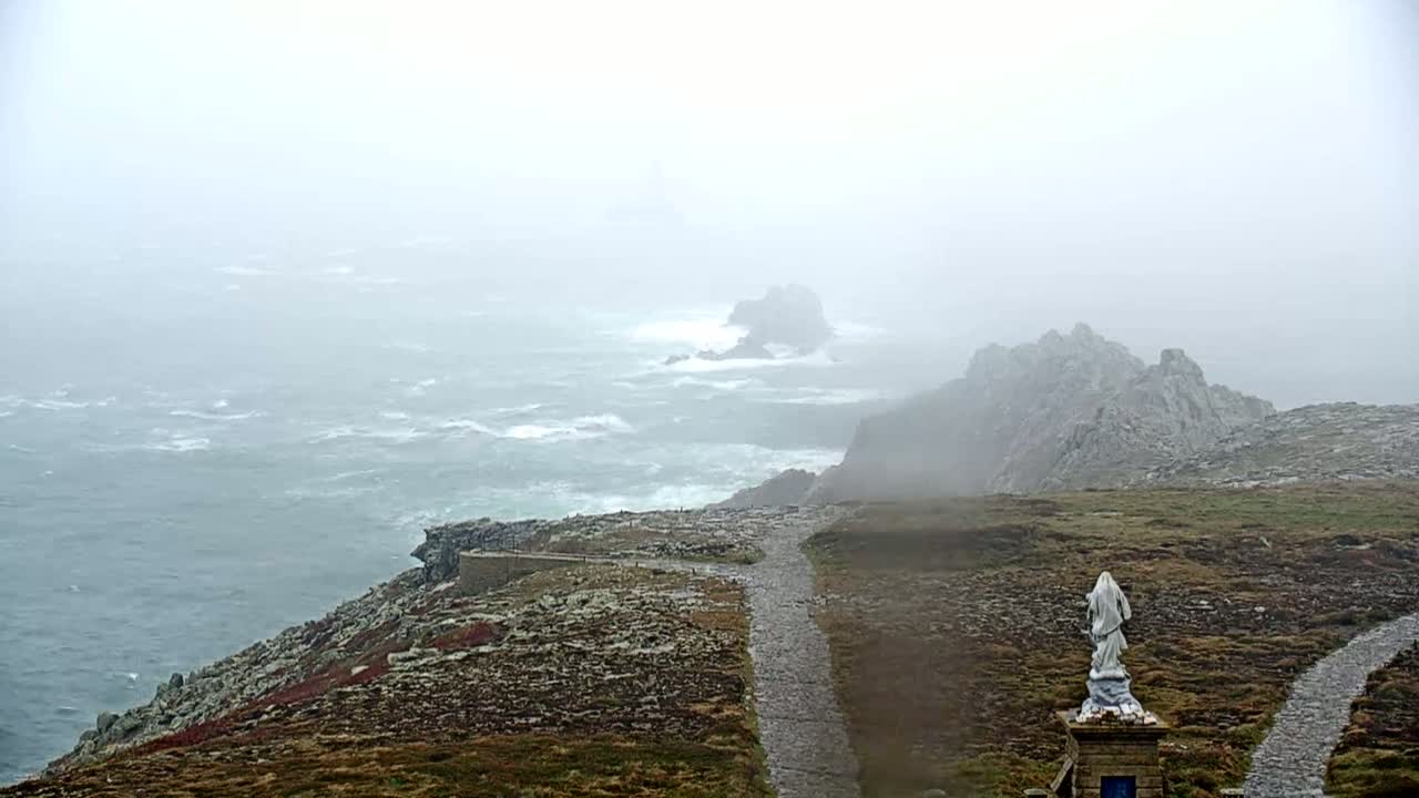 A statue stands atop a rocky coastal path overlooking a turbulent, foggy sea.