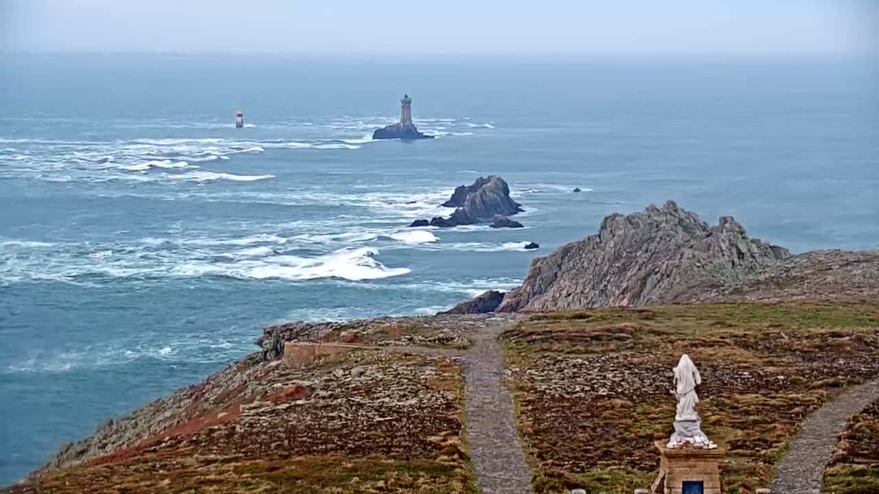 A statue stands on a grassy coastal path overlooking a choppy sea with two lighthouses visible in the distance under an overcast sky.