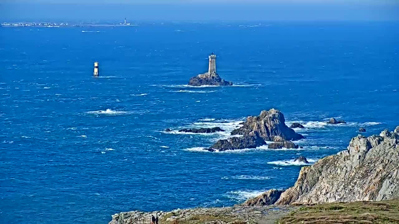 A rocky coastline with a lighthouse on a small island offshore, viewed on a windy, sunny day over a choppy blue sea.