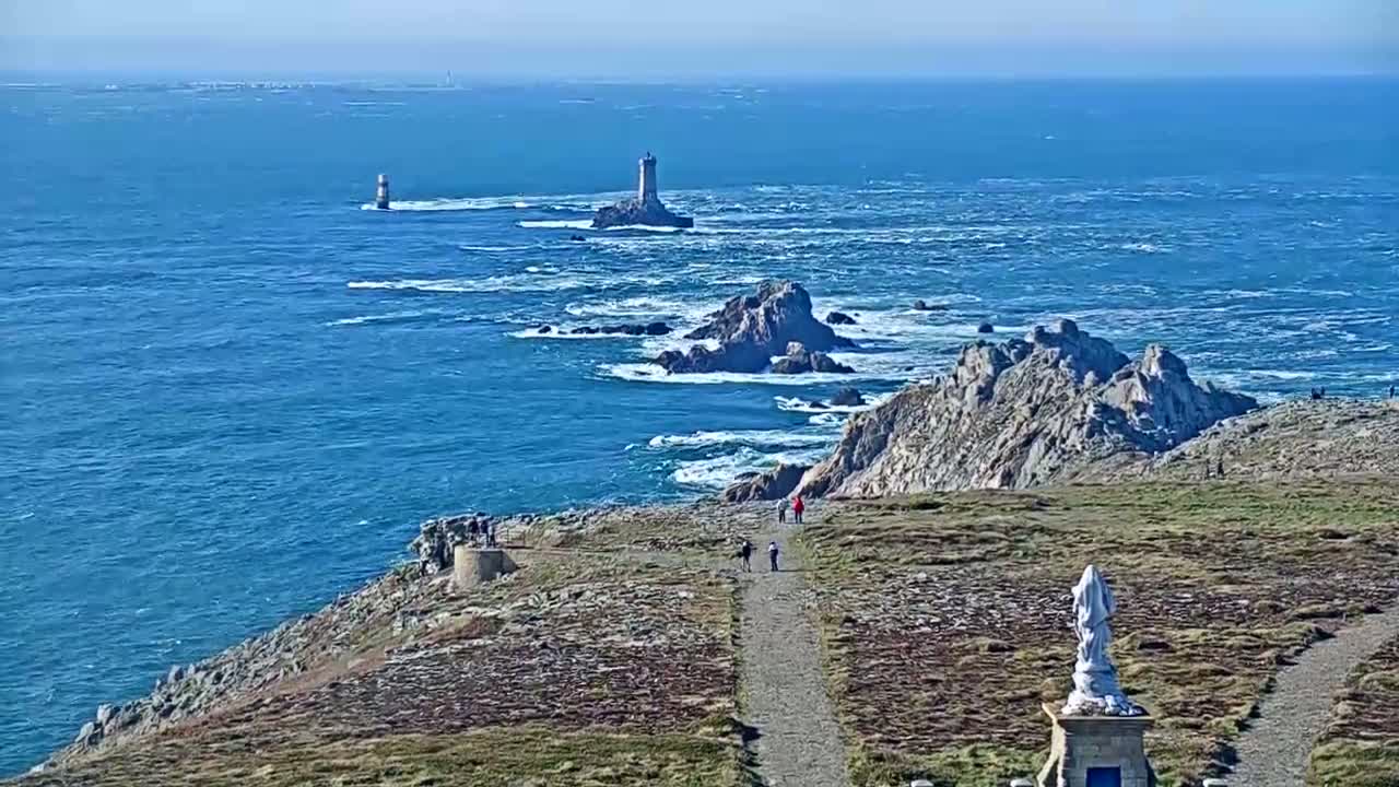 A coastal path overlooks a rocky shoreline and ocean with several lighthouses visible in the distance on a sunny day with a slight wind causing small waves.
