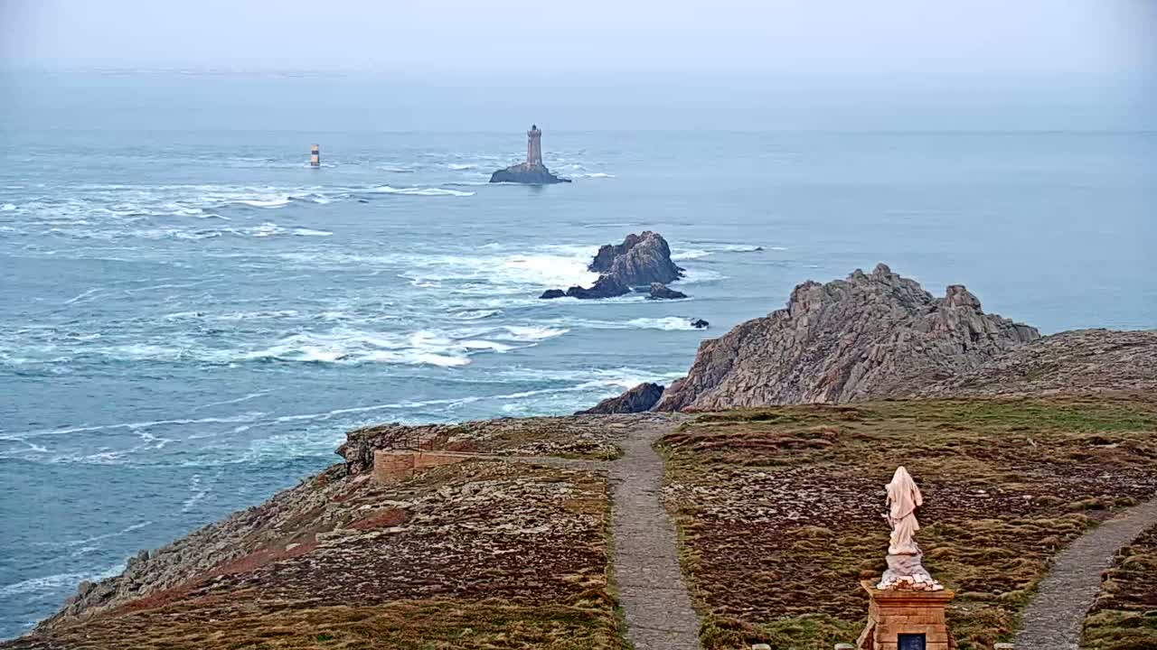 A statue overlooks a rocky coastline and a choppy sea with two lighthouses visible in the distance under an overcast sky.