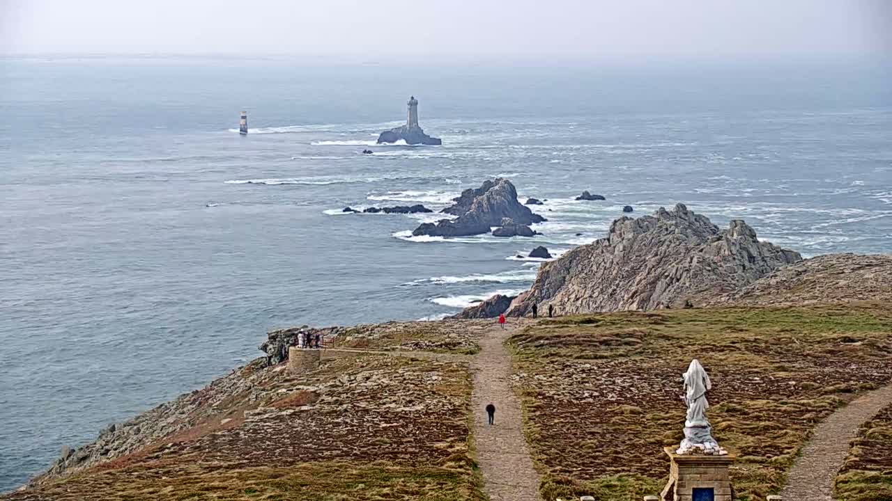 A coastal overlook shows a statue, pathways, and distant lighthouses on rocky islands in a gray, overcast ocean.