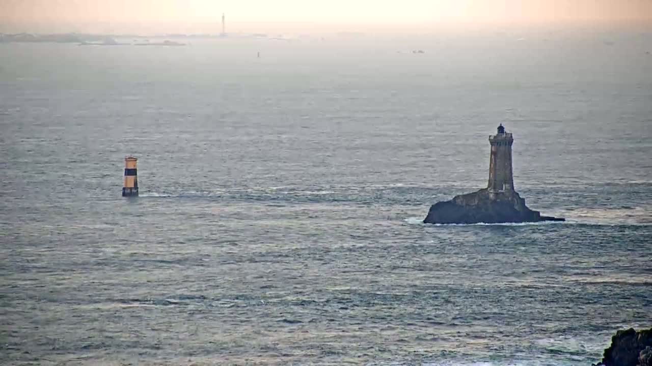 A tall lighthouse sits on a small, rocky island in a hazy, gray sea, with a smaller, striped lighthouse visible in the distance.