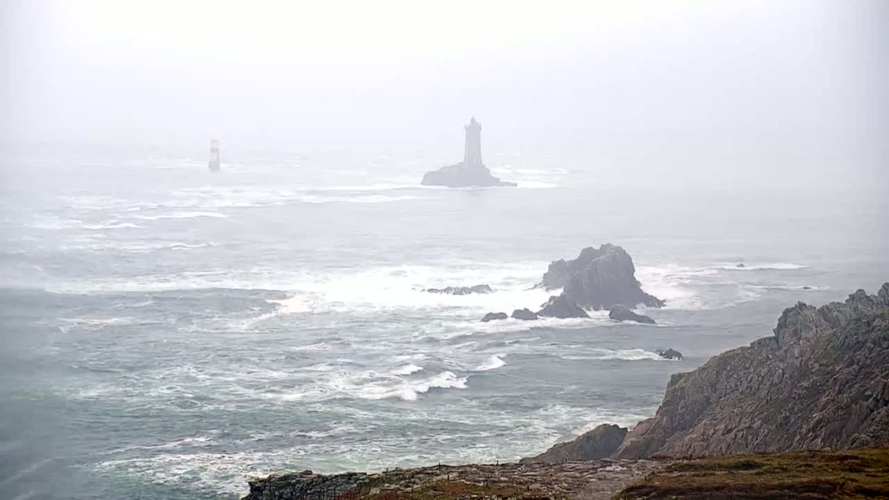 A foggy coastal scene shows a rocky shoreline with crashing waves and two lighthouses visible in the distance.
