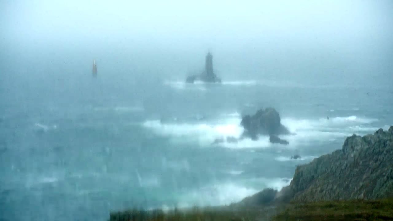 A lighthouse and rocky outcroppings are partially obscured by a heavy mist and large waves in a stormy ocean.