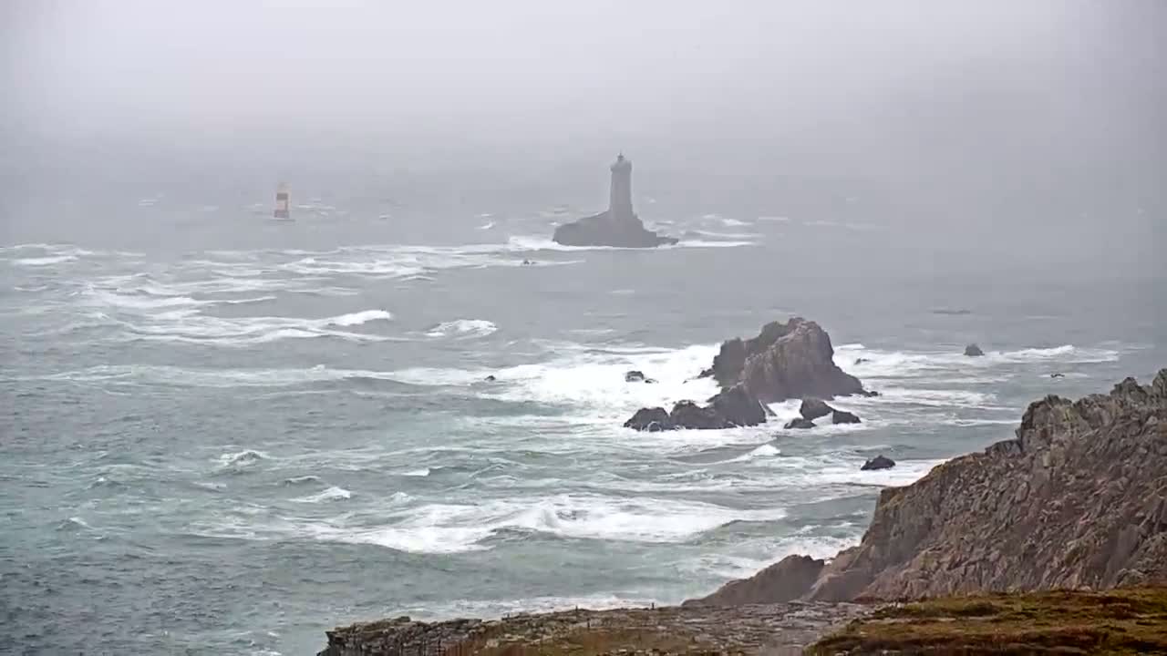 A rocky coastline with two lighthouses visible in the distance is battered by a stormy sea under a foggy sky.