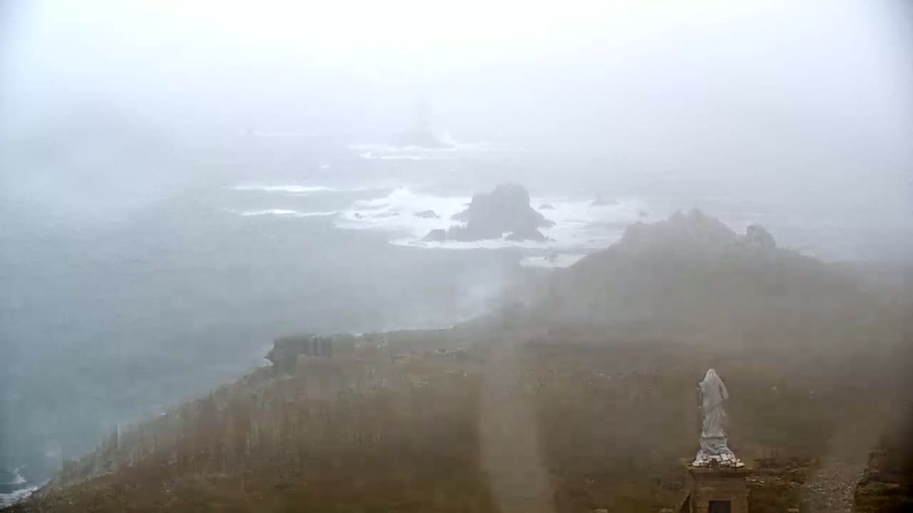 A statue stands atop a rocky outcrop overlooking a foggy, churning ocean with distant rock formations.