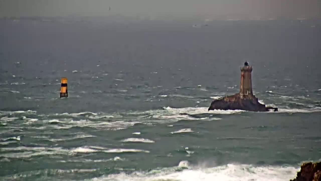 A lighthouse sits on a small, rocky island in a choppy, gray sea, with a navigational marker visible in the distance.