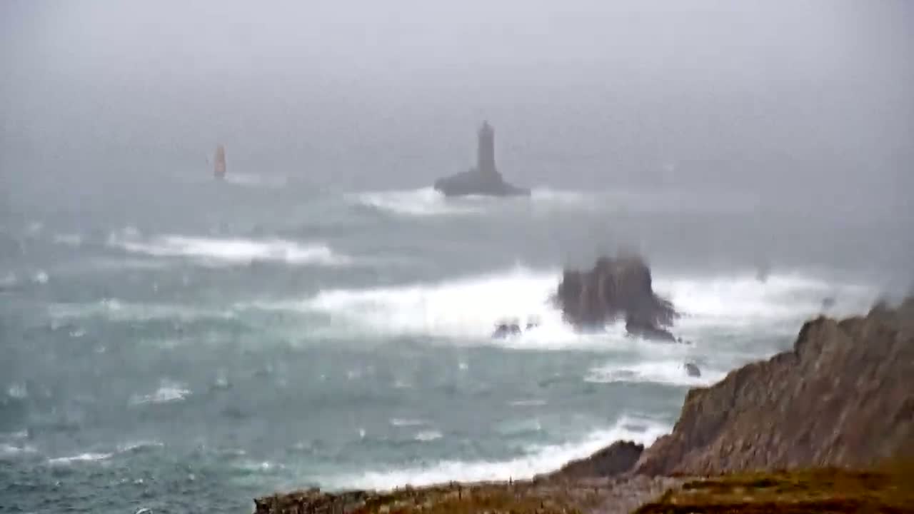 A small sailboat is visible on a rough, stormy sea with large waves crashing against rocky cliffs.