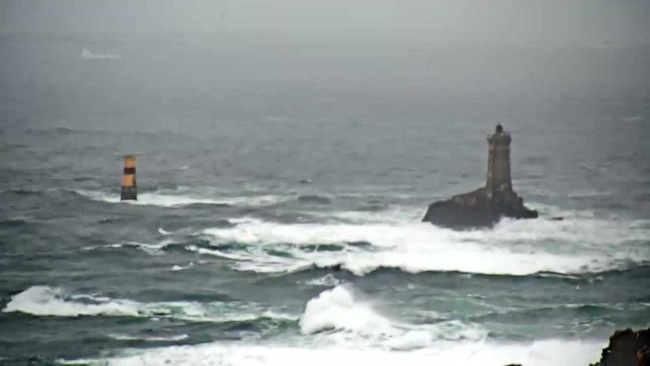A lighthouse sits on a small, rocky island surrounded by rough, choppy waves in an overcast, stormy sea, with a smaller marker buoy visible in the distance.