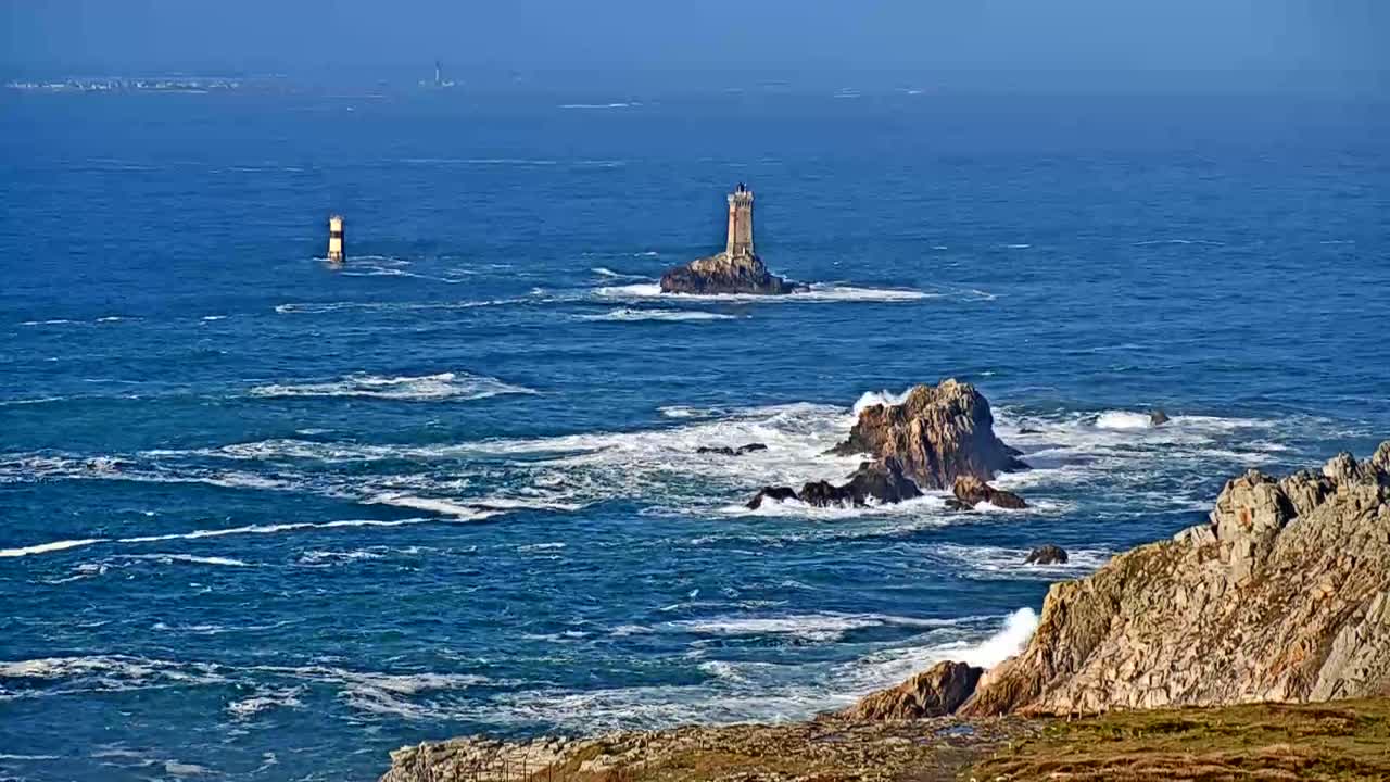 A rocky coastline with crashing waves is visible, featuring two lighthouses on distant islands under a clear, sunny sky.