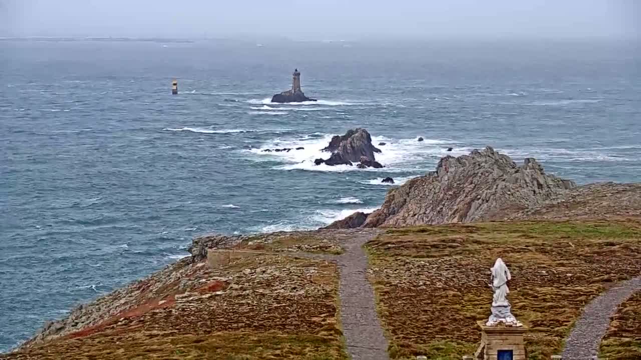 A rocky coastline with a statue, a path, and two lighthouses visible in the distance on a windy, overcast day.
