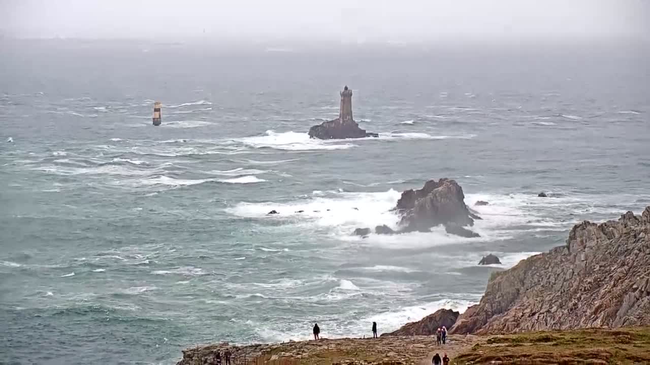 A rocky coastline with a lighthouse on a distant islet is battered by a rough, gray sea under an overcast sky.
