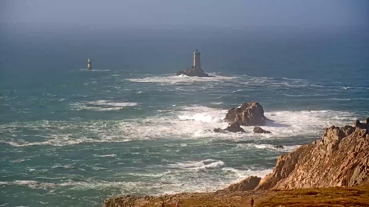 A rocky coastline with two lighthouses visible in the distance, one on a small island, under a hazy, sunny sky with choppy waves crashing against the rocks.