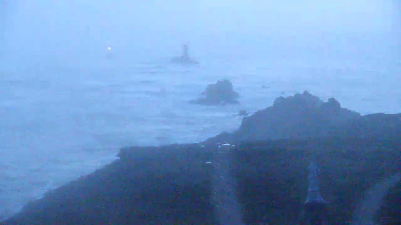 A hazy, stormy seascape shows dark rocky coastlines and distant lighthouses in a blue-gray sea under a foggy sky.