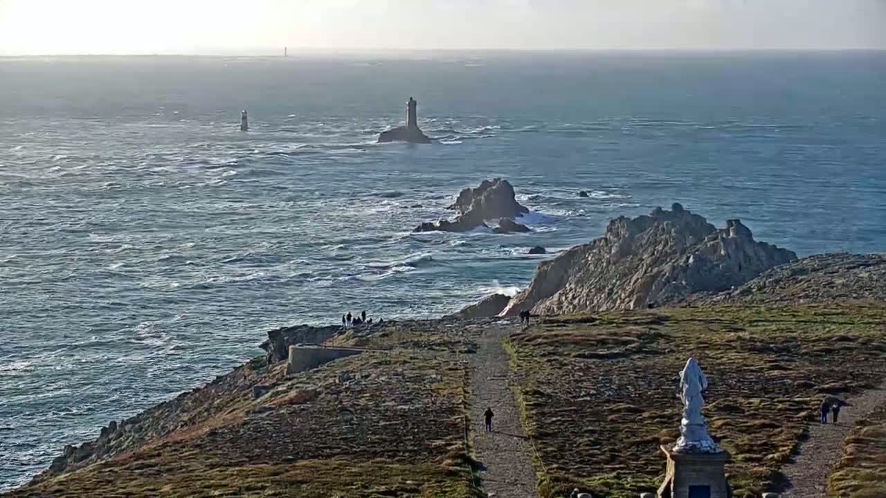 A rocky, grassy coastal area with a statue overlooks a choppy ocean, featuring several distant lighthouses on rocky outcrops under a mostly clear sky.