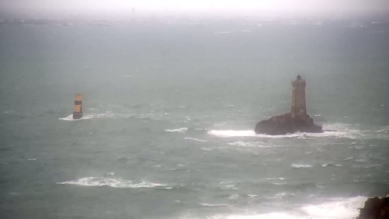 A lighthouse stands on a small, rocky island in a choppy sea during a stormy, overcast day.
