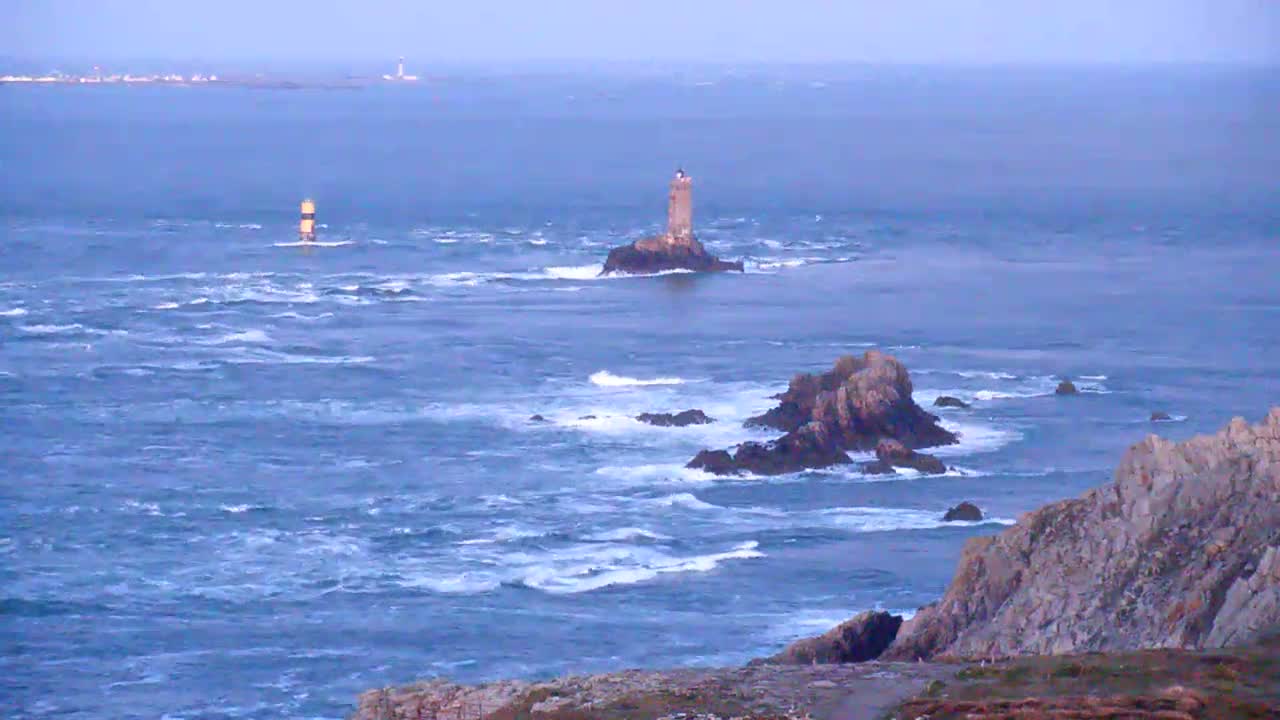 A rocky coastline with two lighthouses visible on distant islands in a choppy sea under a hazy sky.