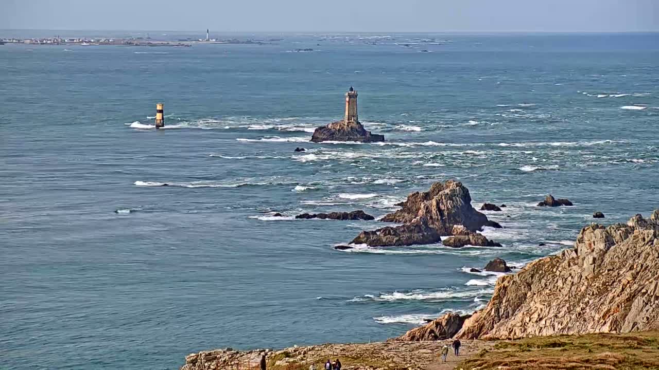 A rocky coastline with a lighthouse on a small island in a choppy ocean under a clear sky.