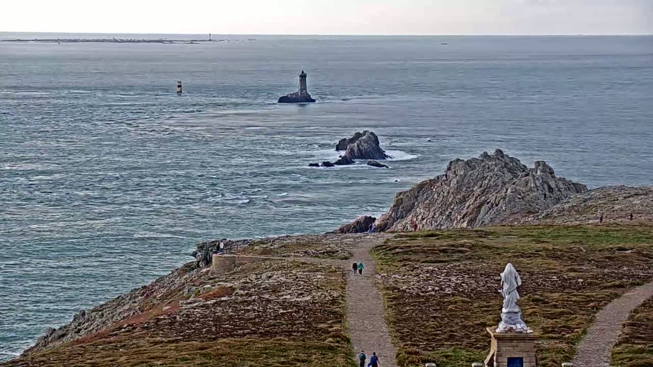 A coastal path winds down a rocky hillside toward a statue, overlooking a choppy sea with a distant lighthouse and several small islands under a cloudy sky.