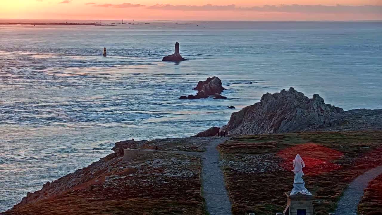 A rocky coastal area at sunset overlooks a body of water with several small islands and lighthouses, featuring a statue at the foreground.