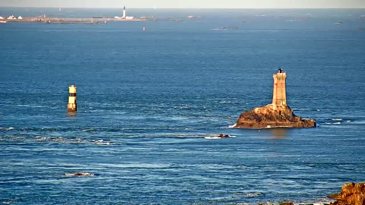 A stone lighthouse sits on a small, rocky island in a calm, blue ocean, with a smaller, striped marker further away.