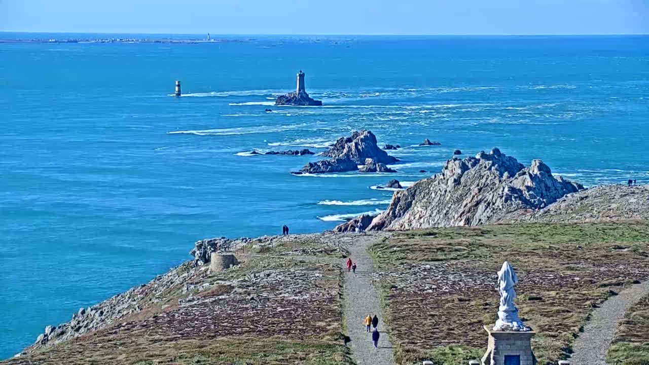 A coastal path winds down a grassy hill towards a statue overlooking a bright blue ocean dotted with rocky islands and lighthouses under sunny skies.