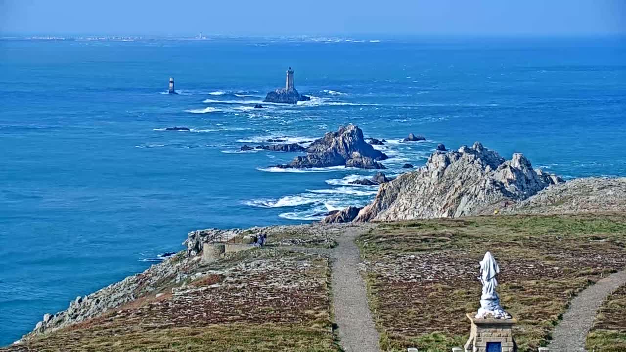A statue overlooks a coastal path and rocky shoreline with crashing waves and distant lighthouses on a clear, sunny day.